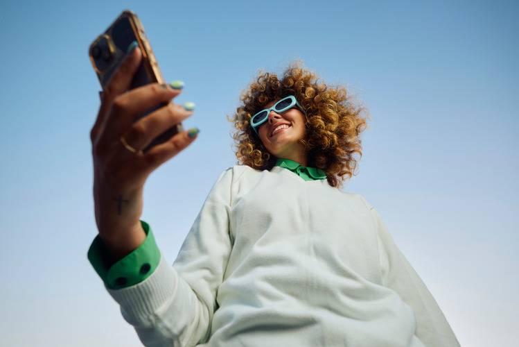 Person med lockigt hår håller upp en smartphone, sedd från en låg vinkel mot en klarblå himmel