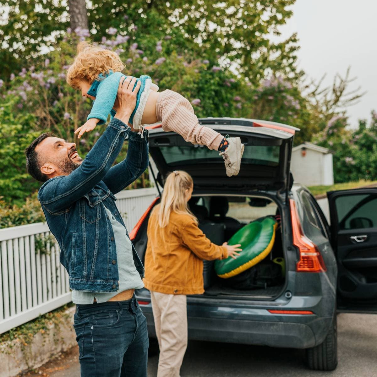 En man som håller upp ett barn ovanför sig och ler. I bakgrunden står en kvinna och packar in saker i en baklucka på en bil.