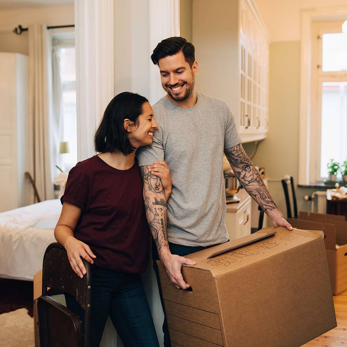 En man bär en flyttkartong i en lägenhet och blir något omkramad av en kvinna som håller i en stol.