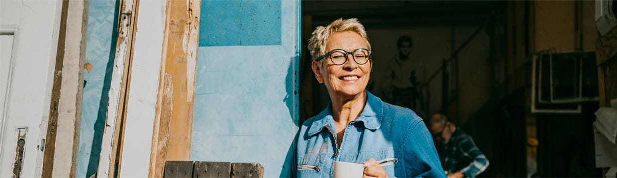 En leende kvinna i kort ljust hår med ljusblå skjorta och glasögon håller i en kopp och lutar sig mot en turkosblå dörr.